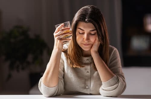 woman sitting with her elbows on a table, holding a glass of alcohol in one hand and resting her chin on the other. She has a hopeless or sad expression on her face.